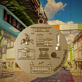 Halskette mit Drehwirbel und Vorderseite eines Schmuckanhängers aus vergoldetem Edelstahl, auf dem Reliefs bekannter Sehenswürdigkeiten der Stadt Wuppertal abgebildet sind. Den Hintergrund bildet eine Ansicht der Stadt Wuppertal.
