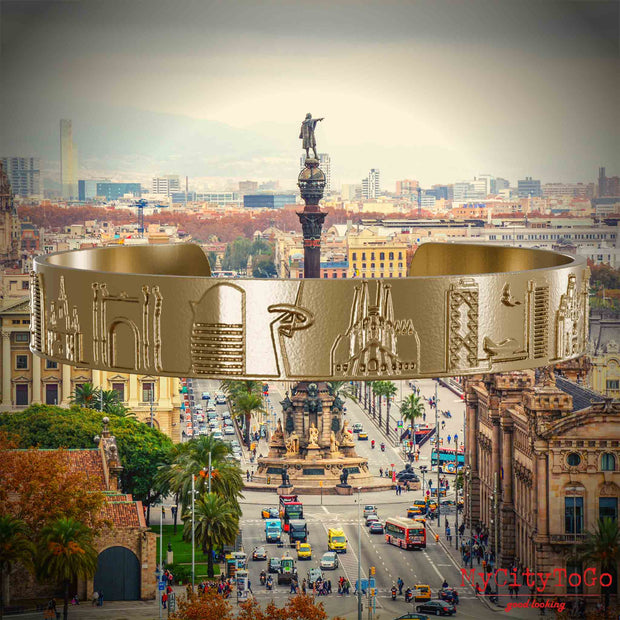 Armreif aus vergoldetem Edelstahl mit Reliefs bekannter Sehenswürdigkeiten der Stadt Barcelona. Den Hintergrund bildet eine Ansicht der Stadt Barcelona.