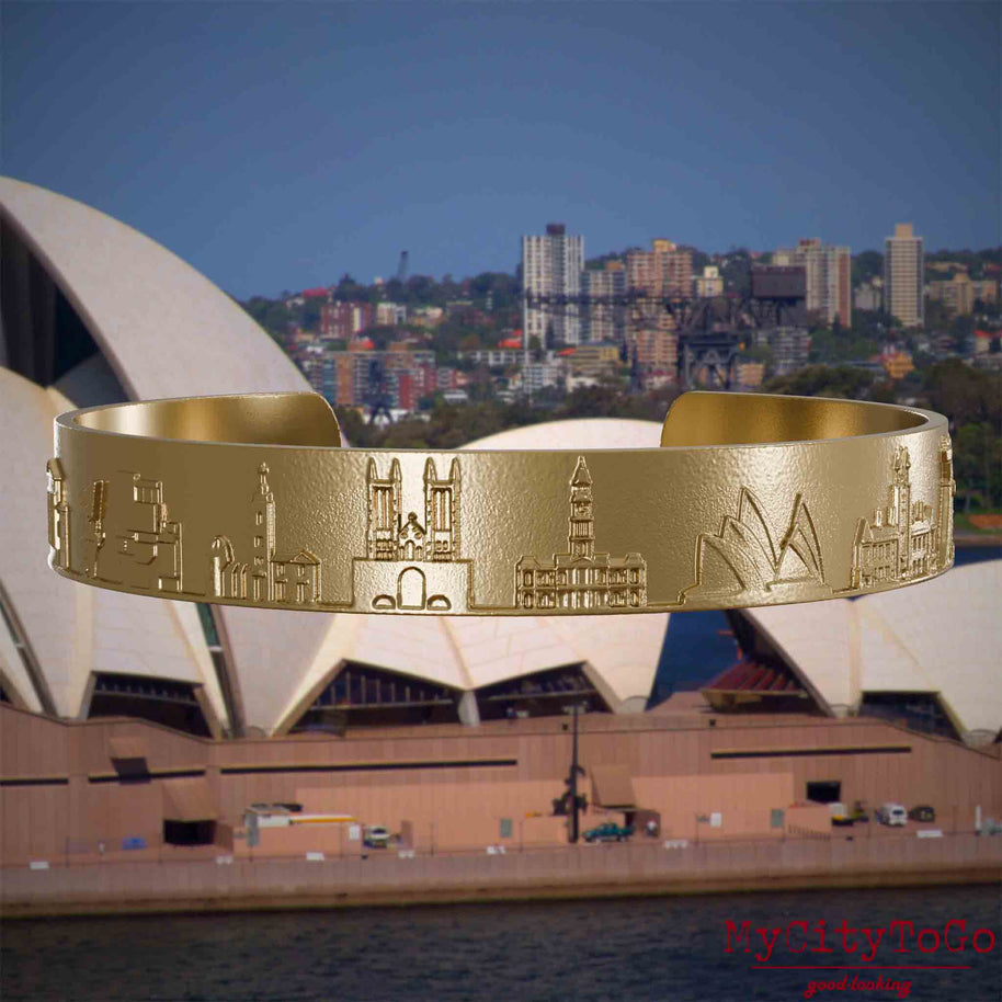 Armreif aus vergoldetem Edelstahl mit Reliefs bekannter Sehenswürdigkeiten der Stadt Sydney. Den Hintergrund bildet eine Ansicht der Stadt Sydney.
