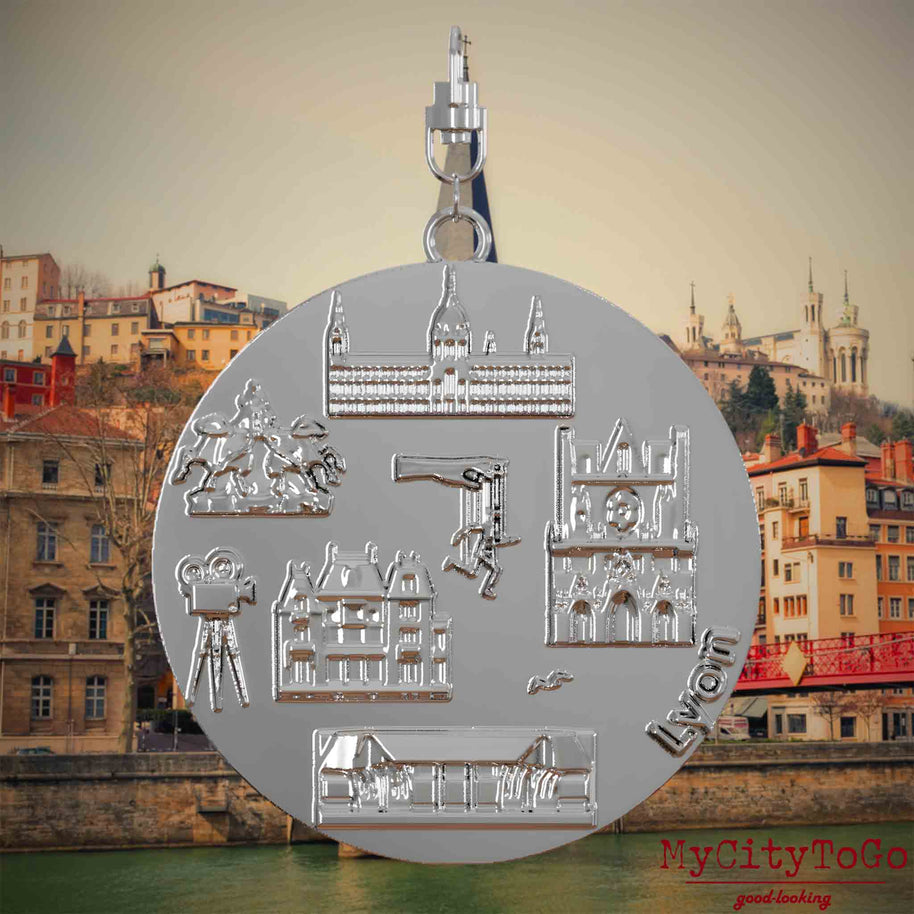 Halskette mit Drehwirbel und Vorderseite eines silbernen Schmuckanhängers, auf dem Reliefs bekannter Sehenswürdigkeiten der Stadt Lyon abgebildet sind. Den Hintergrund bildet eine Ansicht der Stadt Lyon.
