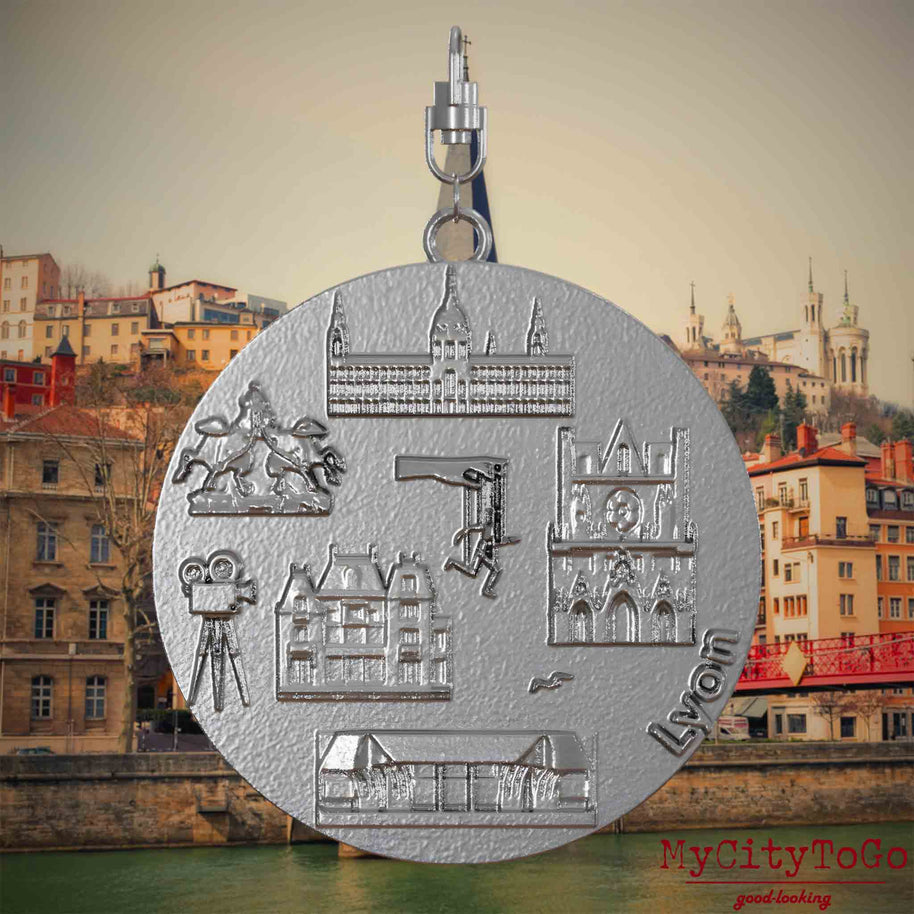 Halskette mit Drehwirbel und Vorderseite eines Schmuckanhängers aus Edelstahl, auf dem Reliefs bekannter Sehenswürdigkeiten der Stadt Lyon abgebildet sind. Den Hintergrund bildet eine Ansicht der Stadt Lyon.
