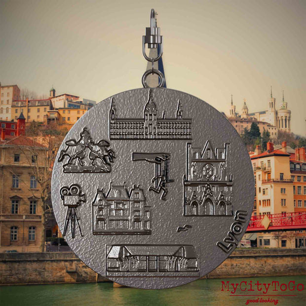 Halskette mit Drehwirbel und Vorderseite eines Schmuckanhängers aus Titan, auf dem Reliefs bekannter Sehenswürdigkeiten der Stadt Lyon abgebildet sind. Den Hintergrund bildet eine Ansicht der Stadt Lyon.
