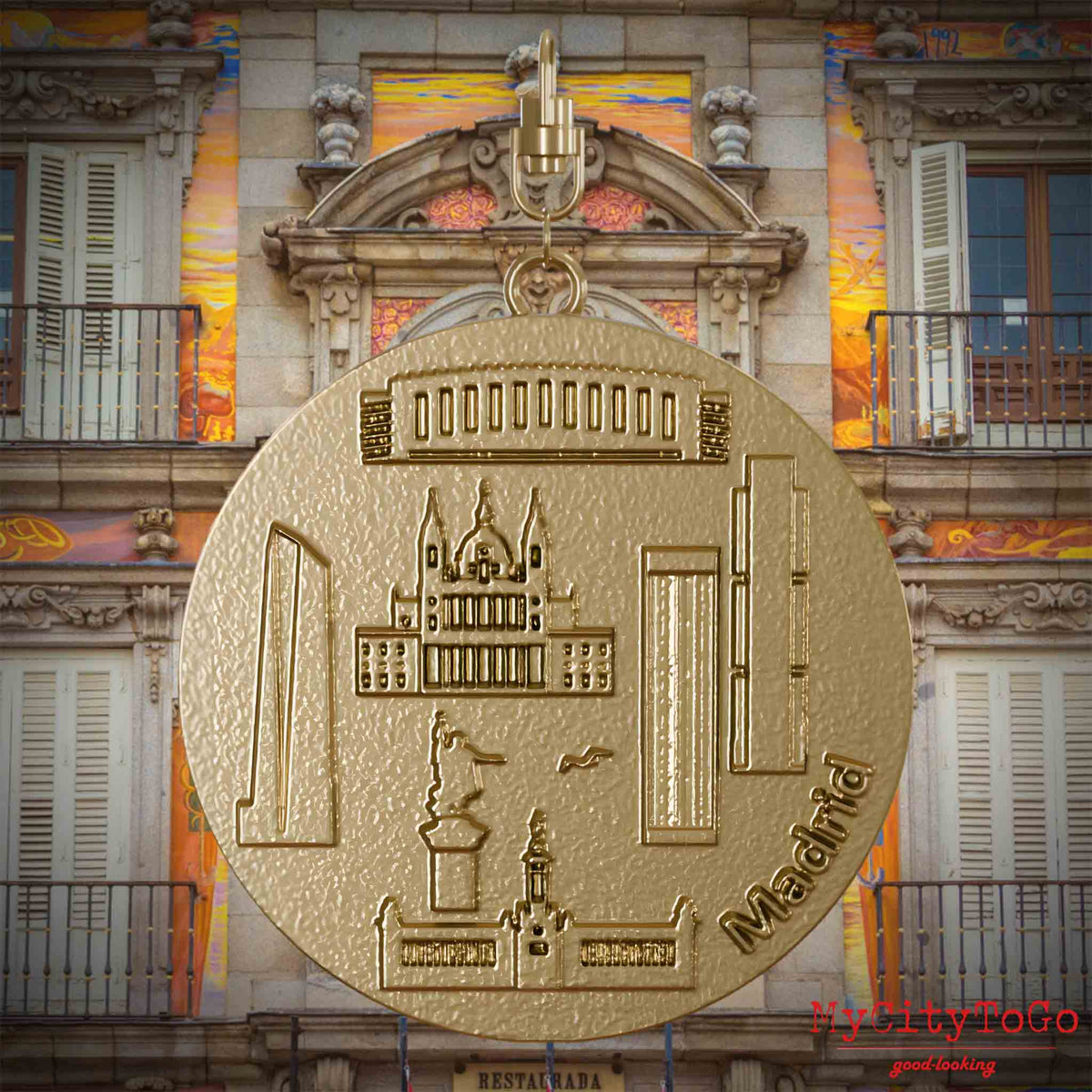 Halskette mit Drehwirbel und Rückseite eines Schmuckanhängers aus vergoldetem Edelstahl, auf dem Reliefs bekannter Sehenswürdigkeiten der Stadt Madrid abgebildet sind. Den Hintergrund bildet eine Ansicht der Stadt Madrid.
