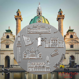 Halskette mit Drehwirbel und Rückseite eines silbernen Schmuckanhängers, auf dem Reliefs bekannter Sehenswürdigkeiten der Stadt Wien abgebildet sind. Den Hintergrund bildet eine Ansicht der Stadt Wien.
