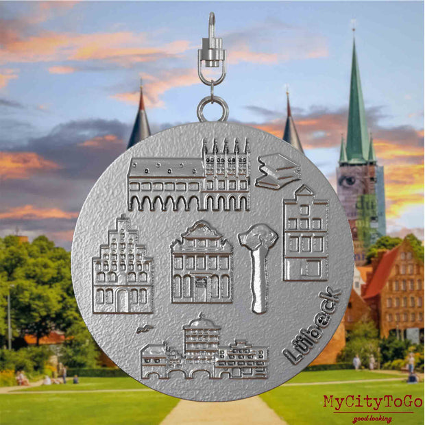 Halskette mit Drehwirbel und Vorderseite eines Schmuckanhängers aus Edelstahl, auf dem Reliefs bekannter Sehenswürdigkeiten der Stadt Luebeck abgebildet sind. Den Hintergrund bildet eine Ansicht der Stadt Luebeck.
