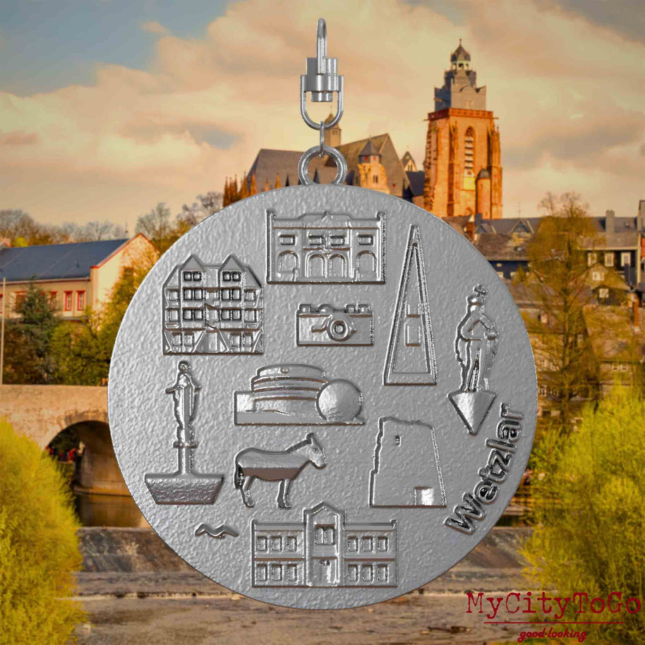 Halskette mit Drehwirbel und Vorderseite eines Schmuckanhängers aus Edelstahl, auf dem Reliefs bekannter Sehenswürdigkeiten der Stadt Wetzlar abgebildet sind. Den Hintergrund bildet eine Ansicht der Stadt Wetzlar.
