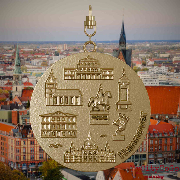 Halskette mit Drehwirbel und Rückseite eines Schmuckanhängers aus vergoldetem Edelstahl, auf dem Reliefs bekannter Sehenswürdigkeiten der Stadt Hannover abgebildet sind. Den Hintergrund bildet eine Ansicht der Stadt Hannover .

