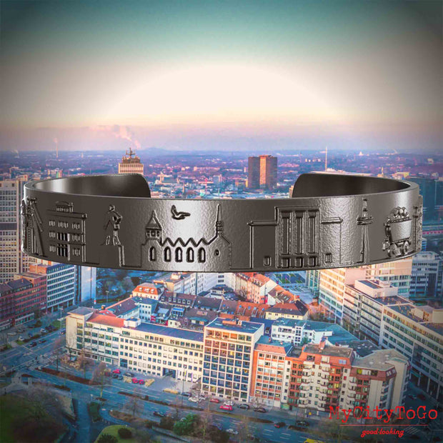 Armreif aus Titan mit Reliefs bekannter Sehenswürdigkeiten der Stadt Essen. Den Hintergrund bildet eine Ansicht der Stadt Essen.