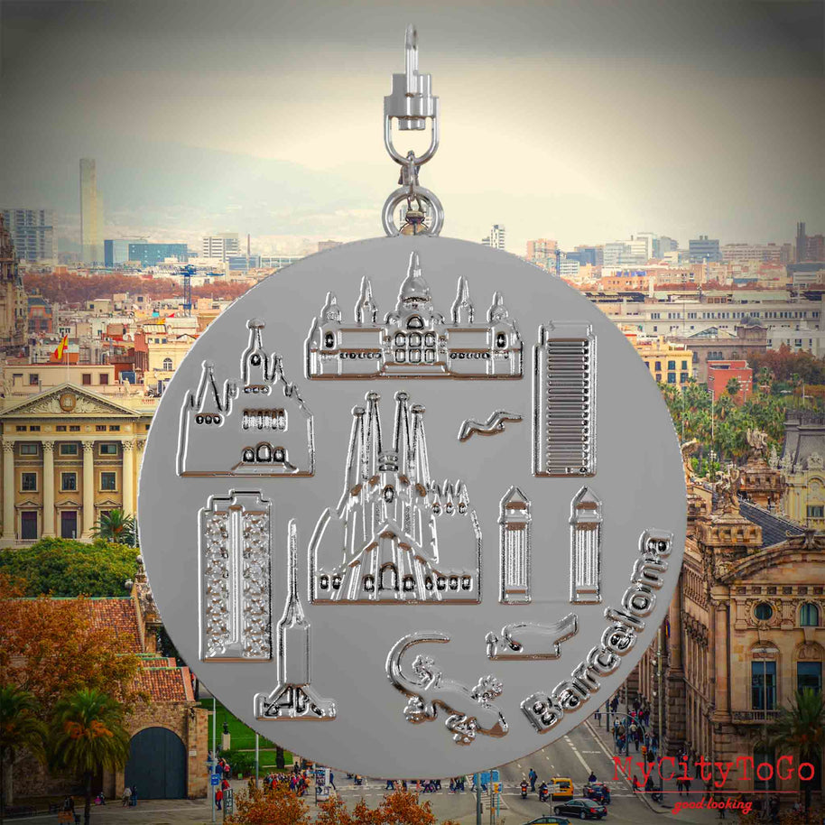 Halskette mit Drehwirbel und Rückseite eines silbernen Schmuckanhängers, auf dem Reliefs bekannter Sehenswürdigkeiten der Stadt Barcelona abgebildet sind. Den Hintergrund bildet eine Ansicht der Stadt Barcelona.
