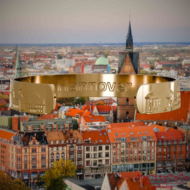Innenseite eines vergoldeten Armreifs mit Reliefs bekannter Sehenswürdigkeiten der Stadt Hannover und einer Gravur des Stadtnamens. Den Hintergrund bildet eine Ansicht der Stadt Hannover.