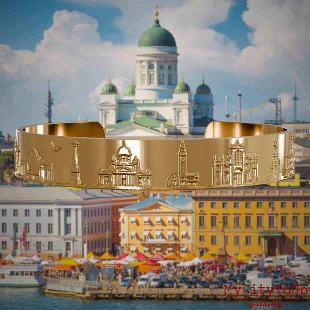Vergoldeter Armreif mit Reliefs bekannter Sehenswürdigkeiten der Stadt Helsinki. Den Hintergrund bildet eine Ansicht der Stadt Helsinki.