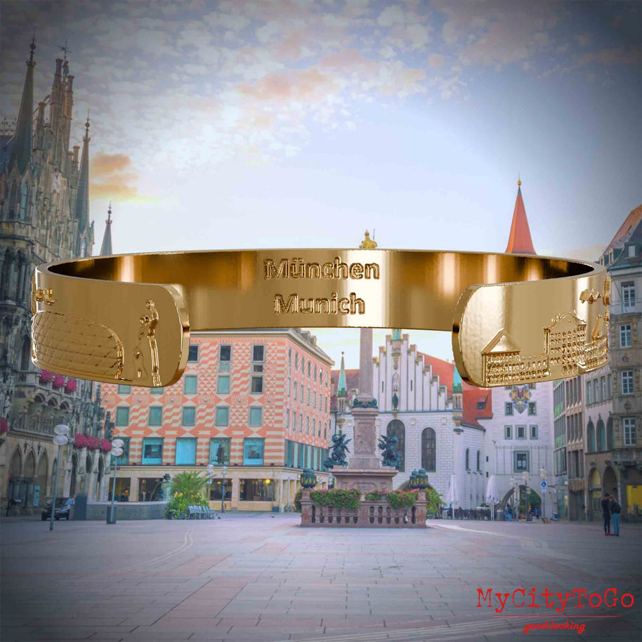 Innenseite eines vergoldeten Armreifs mit Reliefs beliebter Sehenswürdigkeiten der Stadt Müchen und einer Gravur des Stadtnamens in deutsch und englisch. Den Hintergrund bildet eine Ansicht der Stadt Muenchen.
