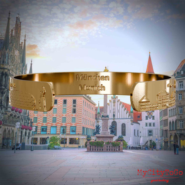 Innenseite eines vergoldeten Armreifs mit Reliefs beliebter Sehenswürdigkeiten der Stadt Müchen und einer Gravur des Stadtnamens in deutsch und englisch. Den Hintergrund bildet eine Ansicht der Stadt Muenchen.