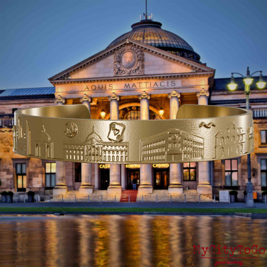 Armreif aus vergoldetem Edelstahl mit Reliefs bekannter Sehenswürdigkeiten der Stadt Wiesbaden. Den Hintergrund bildet eine Ansicht der Stadt Wiesbaden.