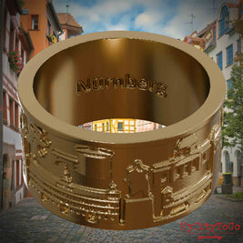 Ring aus vergoldetem Edelstahl mit Reliefs bekannter Sehenswürdigkeiten der Stadt Nuernberg und einer Gravur des Stadtnamens auf der Innenseite. Den Hintergrund bildet eine Ansicht der Stadt  Nuernberg.