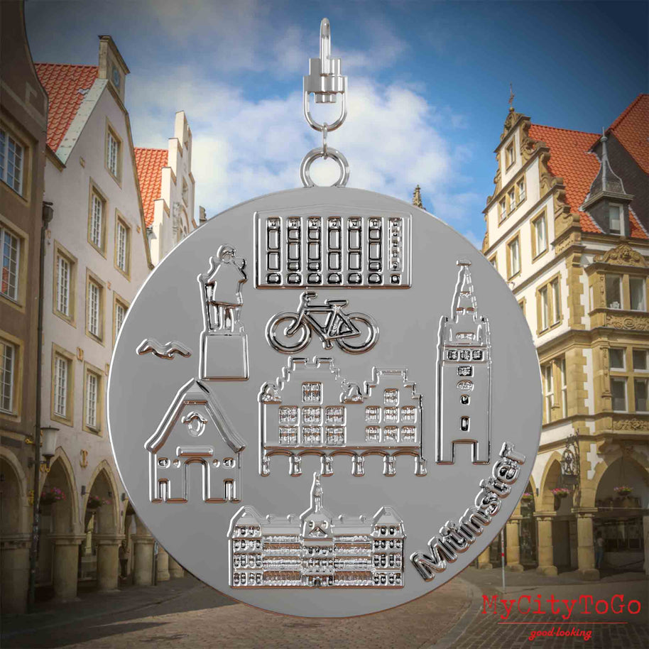 Halskette mit Drehwirbel und Rückseite eines silbernen Schmuckanhängers, auf dem Reliefs bekannter Sehenswürdigkeiten der Stadt Muenster abgebildet sind. Den Hintergrund bildet eine Ansicht der Stadt Muenster.