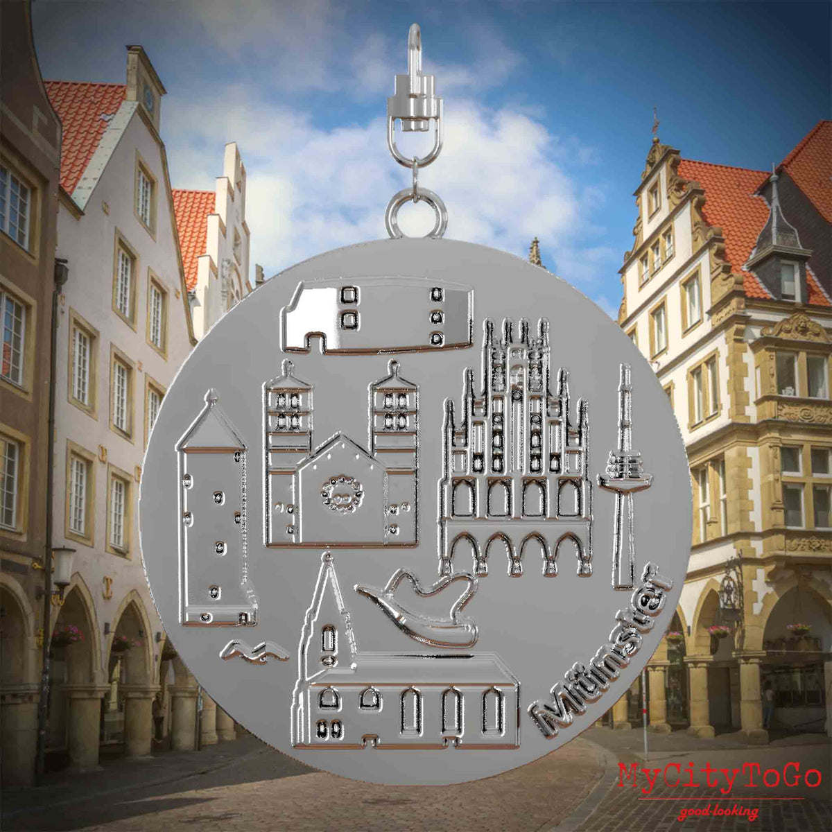 Halskette mit Drehwirbel und Vorderseite eines silbernen Schmuckanhängers, auf dem Reliefs bekannter Sehenswürdigkeiten der Stadt Muenster abgebildet sind. Den Hintergrund bildet eine Ansicht der Stadt Muenster.