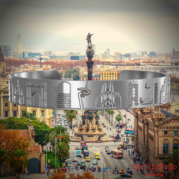 Armreif aus Edelstahl mit Reliefs bekannter Sehenswürdigkeiten der Stadt Barcelona. Den Hintergrund bildet eine Ansicht der Stadt Barcelona.