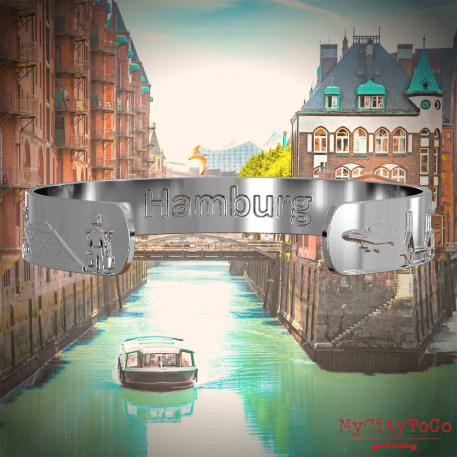 Innenseite eines silbernen Armreifs mit Reliefs bekannter Sehenswürdigkeiten der Stadt Hamburg und einer Gravur des Stadtnamens. Den Hintergrund bildet eine Ansicht der Stadt Hamburg.