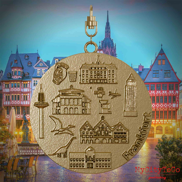 Halskette mit Drehwirbel und Rückseite eines Schmuckanhängers aus vergoldetem Edelstahl, auf dem Reliefs bekannter Sehenswürdigkeiten der Stadt Frankfurt abgebildet sind. Den Hintergrund bildet eine Ansicht der Stadt Frankfurt.