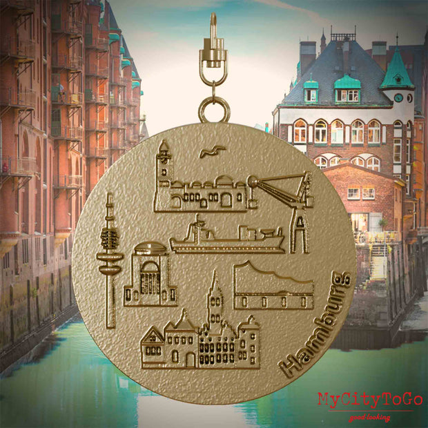 Halskette mit Drehwirbel und Rückseite eines Schmuckanhängers aus vergoldetem Edelstahl, auf dem Reliefs bekannter Sehenswürdigkeiten der Stadt Hamburg abgebildet sind. Den Hintergrund bildet eine Ansicht der Stadt Hamburg.