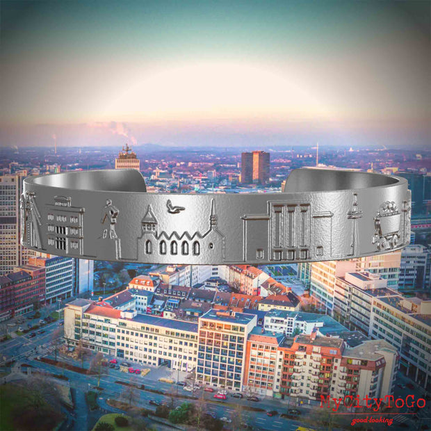 Armreif aus Edelstahl mit Reliefs bekannter Sehenswürdigkeiten der Stadt Essen. Den Hintergrund bildet eine Ansicht der Stadt Essen.