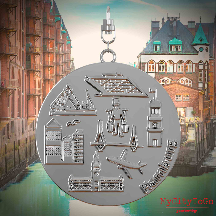 Halskette mit Drehwirbel und Vorderseite eines silbernen Schmuckanhängers, auf dem Reliefs bekannter Sehenswürdigkeiten der Stadt Hamburg abgebildet sind. Den Hintergrund bildet eine Ansicht der Stadt Hamburg.