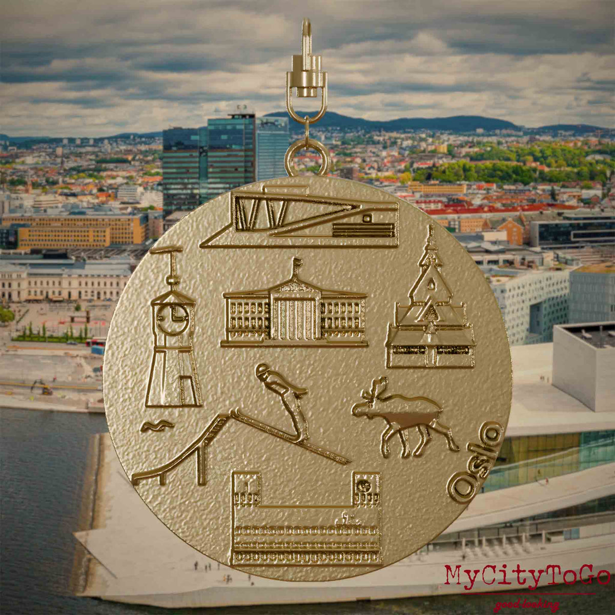 Halskette mit Drehwirbel und Vorderseite eines Schmuckanhängers aus vergoldetem Edelstahl, auf dem Reliefs bekannter Sehenswürdigkeiten der Stadt Oslo abgebildet sind. Den Hintergrund bildet eine Ansicht der Stadt Oslo.
