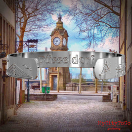 Innenseite eines silbernen Armreifs mit Reliefs bekannter Sehenswürdigkeiten der Stadt Duesseldorf und einer Gravur des Stadtnamens. Den Hintergrund bildet eine Ansicht der Stadt Duesseldorf.