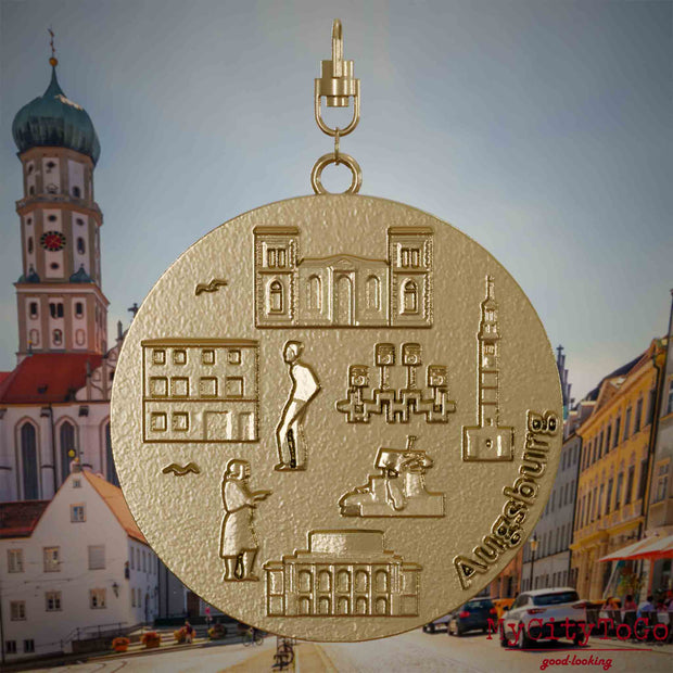 Halskette mit Drehwirbel und Vorderseite eines Schmuckanhängers aus vergoldetem Edelstahl, auf dem Reliefs bekannter Sehenswürdigkeiten der Stadt Augsburg abgebildet sind. Den Hintergrund bildet eine Ansicht der Stadt Augsburg.
