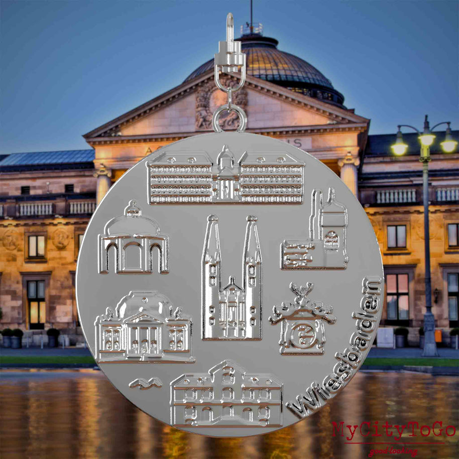 Halskette mit Drehwirbel und Rückseite eines silbernen Schmuckanhängers, auf dem Reliefs bekannter Sehenswürdigkeiten der Stadt Wiesbaden abgebildet sind. Den Hintergrund bildet eine Ansicht der Stadt Wiesbaden.
