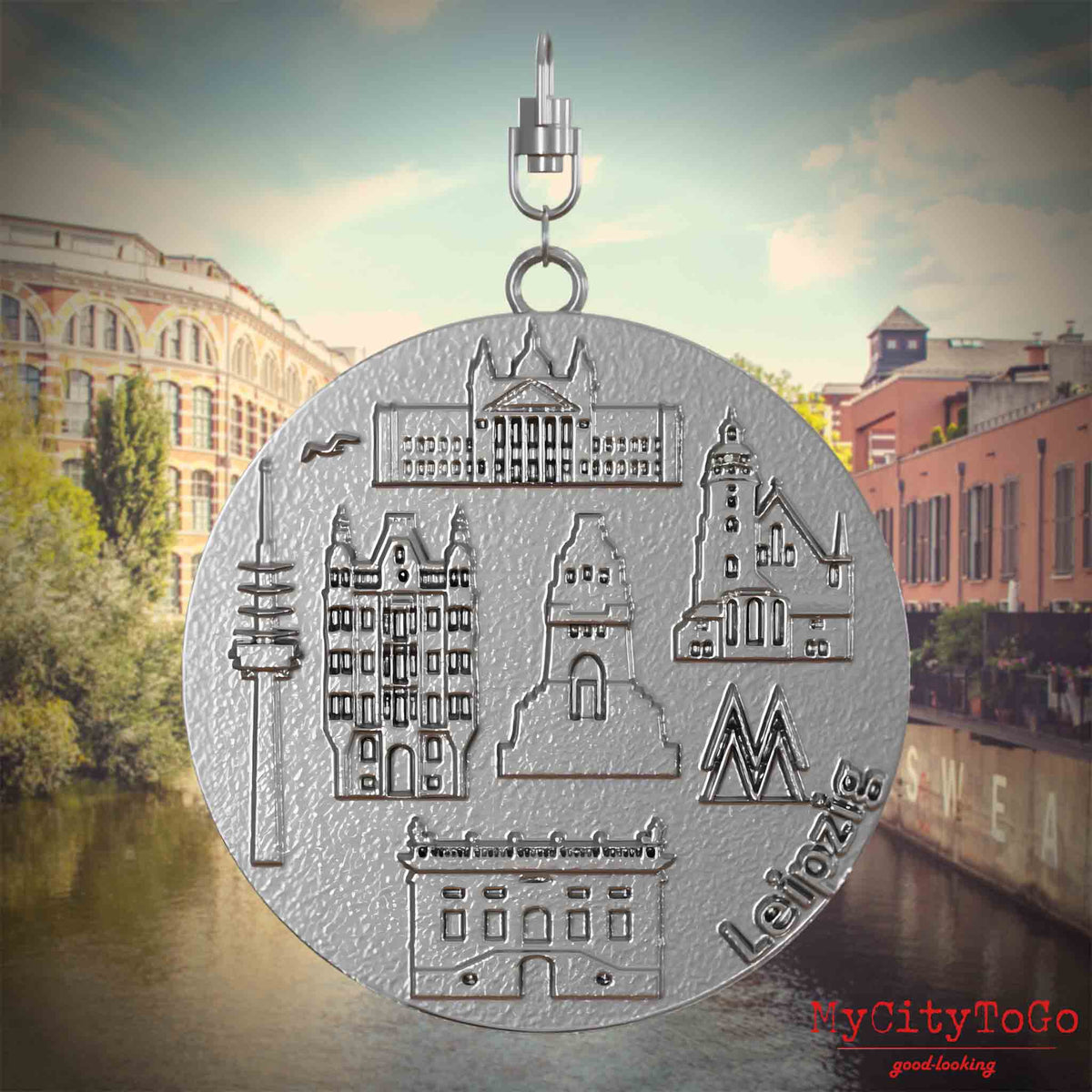 Halskette mit Drehwirbel und Vorderseite eines Schmuckanhänger , auf dem Reliefs bekannter Sehenswürdigkeiten der Stadt Leipzig abgebildet sind. Den Hintergrund bildet eine Ansicht der Stadt Leipzig.
