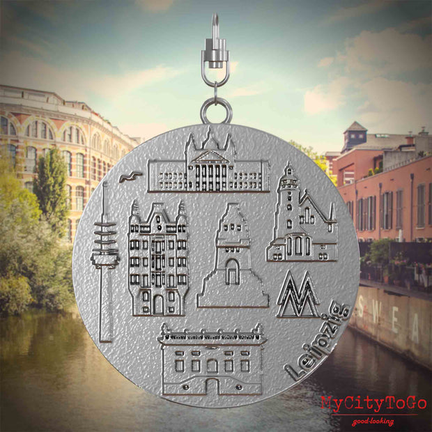Halskette mit Drehwirbel und Vorderseite eines Schmuckanhänger , auf dem Reliefs bekannter Sehenswürdigkeiten der Stadt Leipzig abgebildet sind. Den Hintergrund bildet eine Ansicht der Stadt Leipzig.