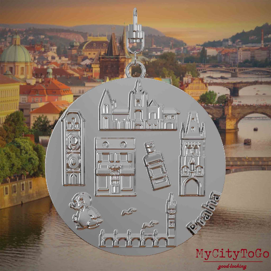 Halskette mit Drehwirbel und Vorderseite eines silbernen Schmuckanhängers, auf dem Reliefs bekannter Sehenswürdigkeiten der Stadt Prag abgebildet sind. Den Hintergrund bildet eine Ansicht der Stadt Prag.
