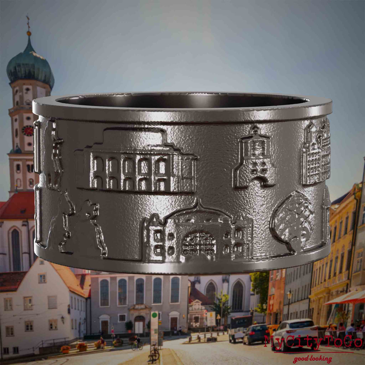 Ring aus Titan mit Reliefs bekannter Sehenswürdigkeiten der Stadt Augsburg. Den Hintergrund bildet eine Ansicht der Stadt Augsburg.