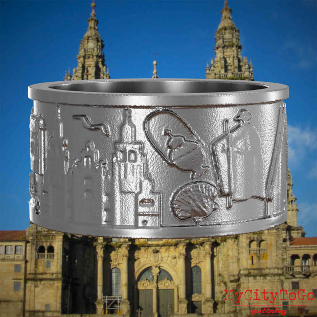 Ring aus Edelstahl mit Reliefs bekannter Sehenswürdigkeiten und Wahrzeichen auf dem Jakobsweg. Den Hintergrund bildet die Kathedrale von Santiago de Compostela.