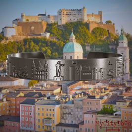 Armreif aus Titan mit Reliefs bekannter Sehenswürdigkeiten der Stadt Salzburg. Den Hintergrund bildet eine Ansicht der Stadt Salzburg.