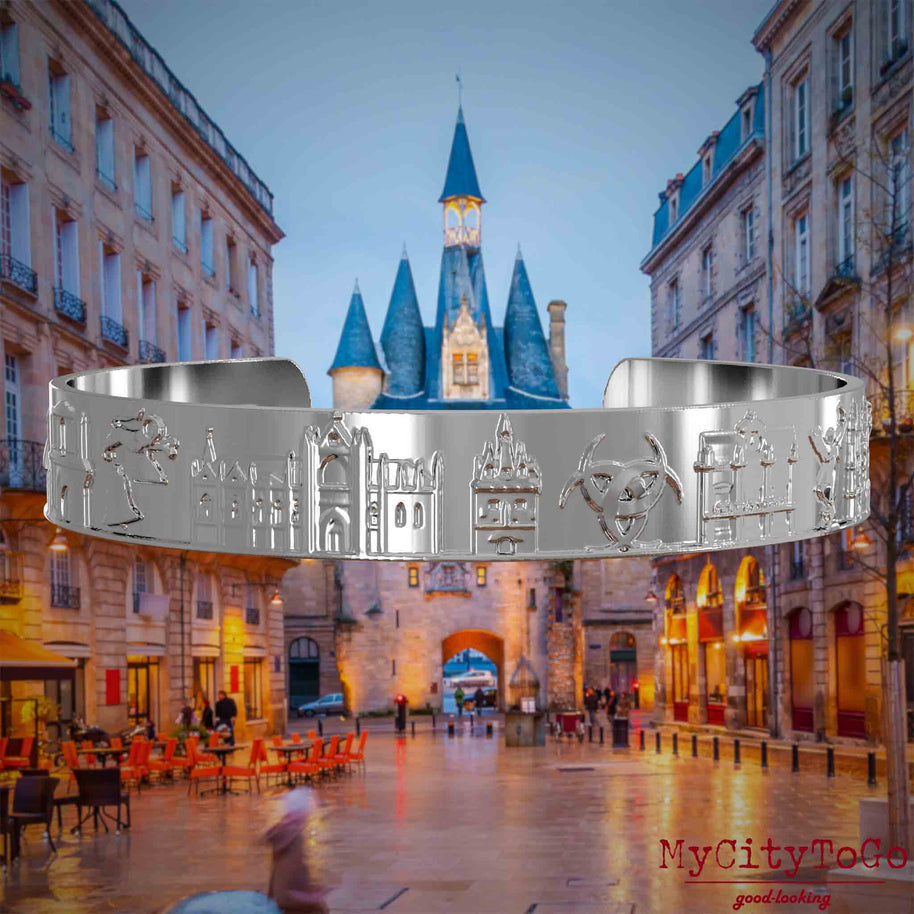 Versilberter Armreif mit Reliefs bekannter Sehenswürdigkeiten der Stadt Bordeaux. Den Hintergrund bildet eine Ansicht der Stadt Bordeaux.