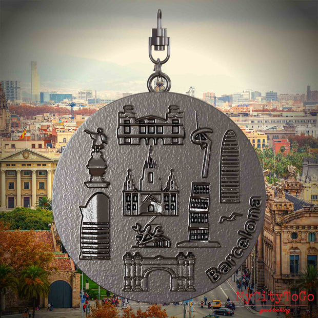 Halskette mit Drehwirbel und Vorderseite eines Schmuckanhängers aus Titan, auf dem Reliefs bekannter Sehenswürdigkeiten der Stadt Barcelona abgebildet sind. Den Hintergrund bildet eine Ansicht der Stadt Barcelona.
