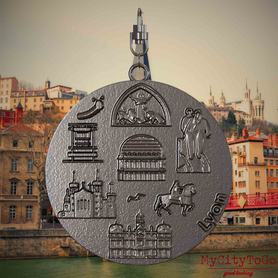 Halskette mit Drehwirbel und Rückseite eines Schmuckanhängers aus Titan, auf dem Reliefs bekannter Sehenswürdigkeiten der Stadt Lyon abgebildet sind. Den Hintergrund bildet eine Ansicht der Stadt Lyon.
