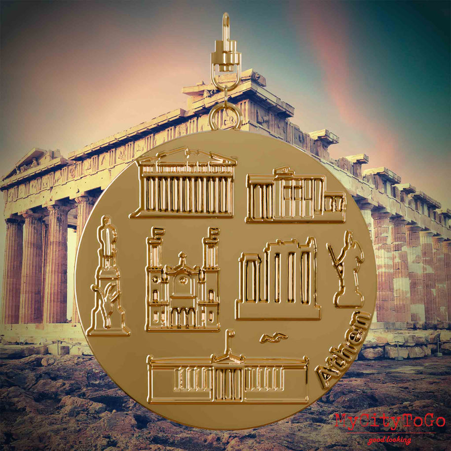 Halskette mit Drehwirbel und Vorderseite eines vergoldeten Schmuckanhängers, auf dem Reliefs bekannter Sehenswürdigkeiten der Stadt Athen abgebildet sind. Den Hintergrund bildet eine Ansicht der Stadt Athen.
