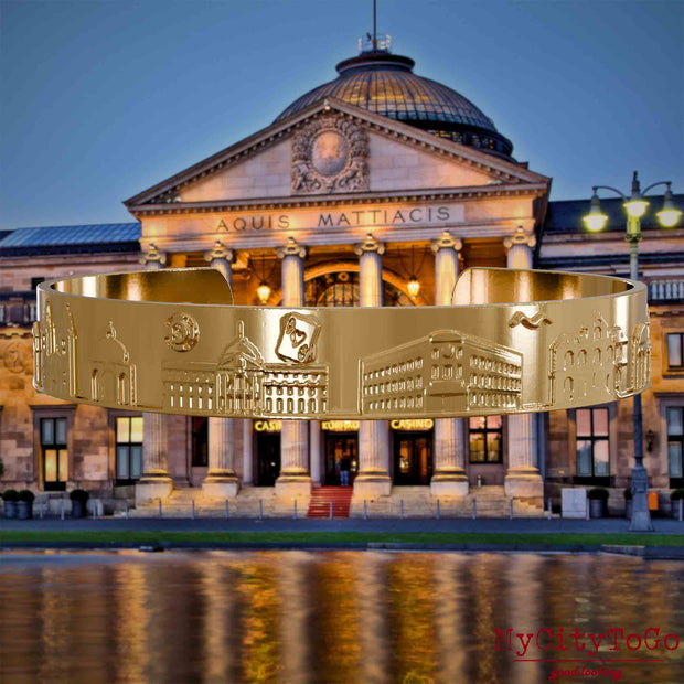 Vergoldeter Armreif mit Reliefs bekannter Sehenswürdigkeiten der Stadt Wiesbaden. Den Hintergrund bildet eine Ansicht der Stadt Wiesbaden.
