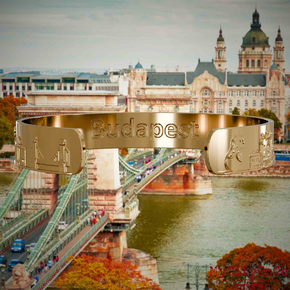 Innenseite eines vergoldeten Armreifs mit Reliefs bekannter Sehenswürdigkeiten der Stadt Budapest und einer Gravur des Stadtnamens. Den Hintergrund bildet eine Ansicht der Stadt Budapest.