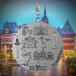 Halskette mit Drehwirbel und Rückseite eines Schmuckanhängers aus Edelstahl, auf dem Reliefs bekannter Sehenswürdigkeiten der Stadt Frankfurt abgebildet sind. Den Hintergrund bildet eine Ansicht der Stadt Frankfurt.