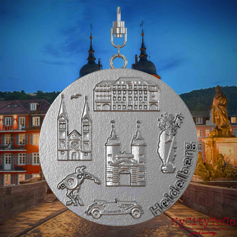 Halskette mit Drehwirbel und Rückseite eines Schmuckanhängers aus Edelstahl, auf dem Reliefs bekannter Sehenswürdigkeiten der Stadt Heidelberg abgebildet sind. Den Hintergrund bildet eine Ansicht der Stadt Heidelberg .
