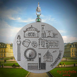 Halskette mit Drehwirbel und Rückseite eines Schmuckanhängers aus Edelstahl, auf dem Reliefs bekannter Sehenswürdigkeiten der Stadt Stuttgart abgebildet sind. Den Hintergrund bildet eine Ansicht der Stadt Stuttgart.
