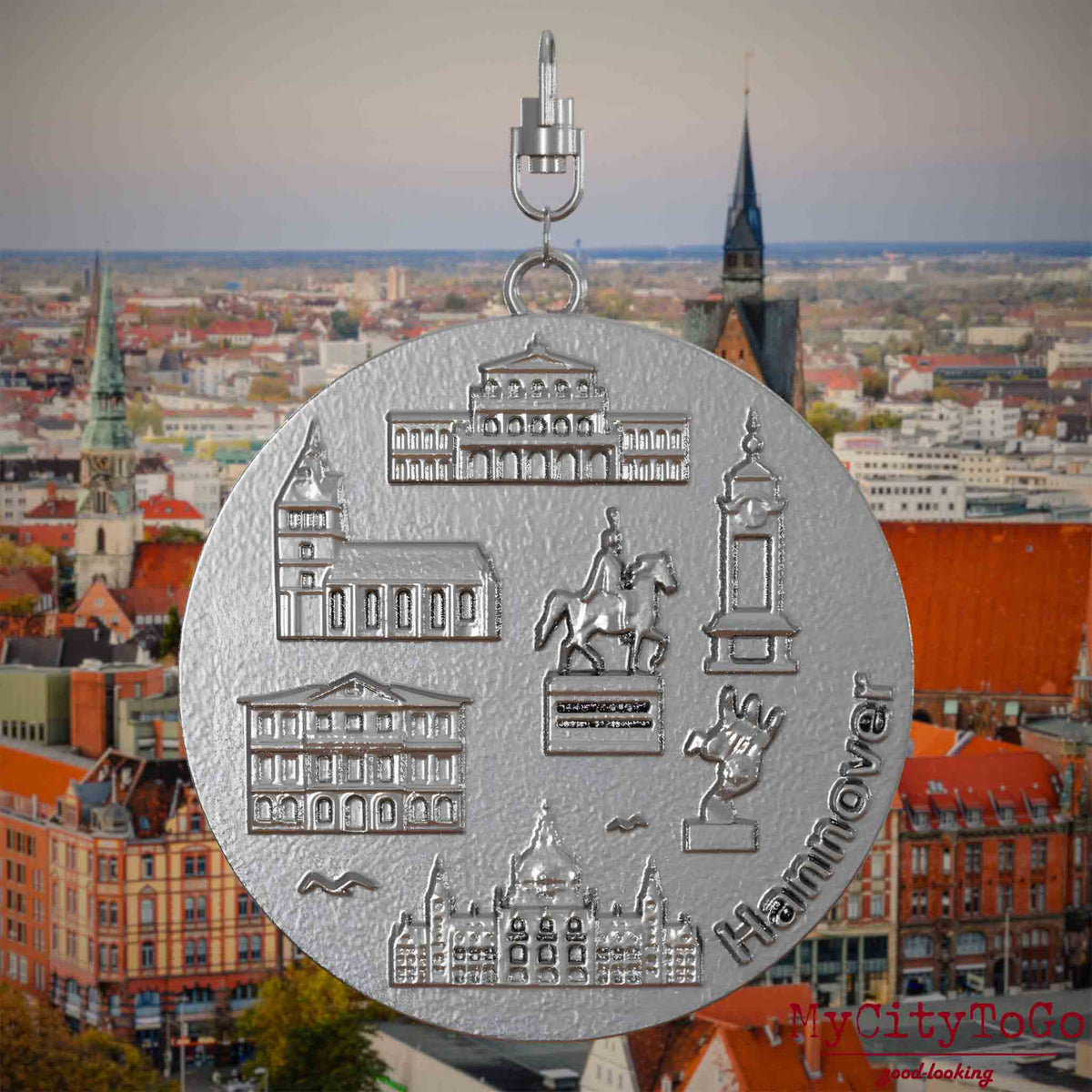 Halskette mit Drehwirbel und Rückseite eines Schmuckanhängers aus Edelstahl, auf dem Reliefs bekannter Sehenswürdigkeiten der Stadt Hannover abgebildet sind. Den Hintergrund bildet eine Ansicht der Stadt Hannover .
