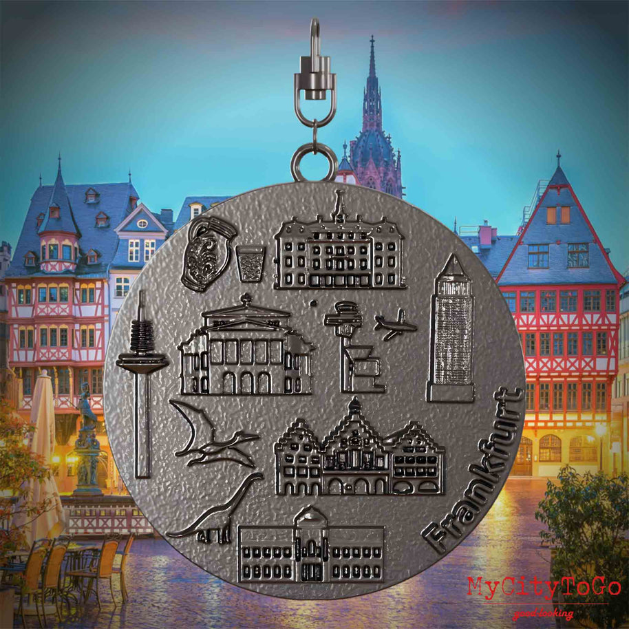 Halskette mit Drehwirbel und Rückseite eines Schmuckanhängers aus Titan, auf dem Reliefs bekannter Sehenswürdigkeiten der Stadt Frankfurt abgebildet sind. Den Hintergrund bildet eine Ansicht der Stadt Frankfurt.
