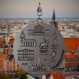Halskette mit Drehwirbel und Rückseite eines Schmuckanhängers aus Titan, auf dem Reliefs bekannter Sehenswürdigkeiten der Stadt Hannover abgebildet sind. Den Hintergrund bildet eine Ansicht der Stadt Hannover .
