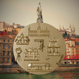 Halskette mit Drehwirbel und Vorderseite eines Schmuckanhängers aus vergoldetem Edelstahl, auf dem Reliefs bekannter Sehenswürdigkeiten der Stadt Lyon abgebildet sind. Den Hintergrund bildet eine Ansicht der Stadt Lyon.
